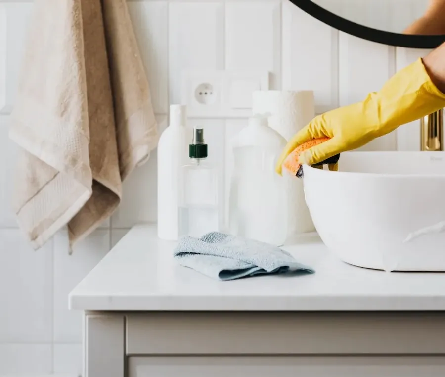 Scrubbing white bathroom sink with yellow gloves and orange sponge